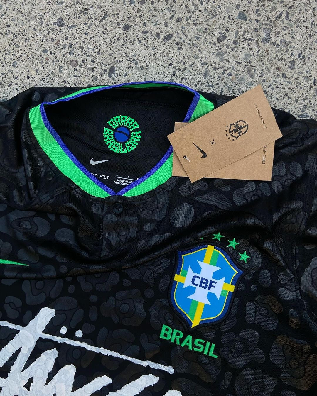 Close-up of a black Brazil x Stussy soccer jersey. The collar features a green and blue stripe with the Nike and CBF logos. The jersey&